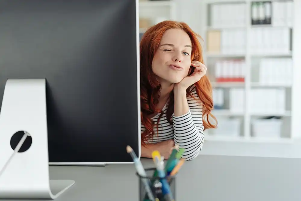 Eine Person lehnt sich augenzwinkernd an einem Schreibtisch neben einem Monitor an (Symbolbild).